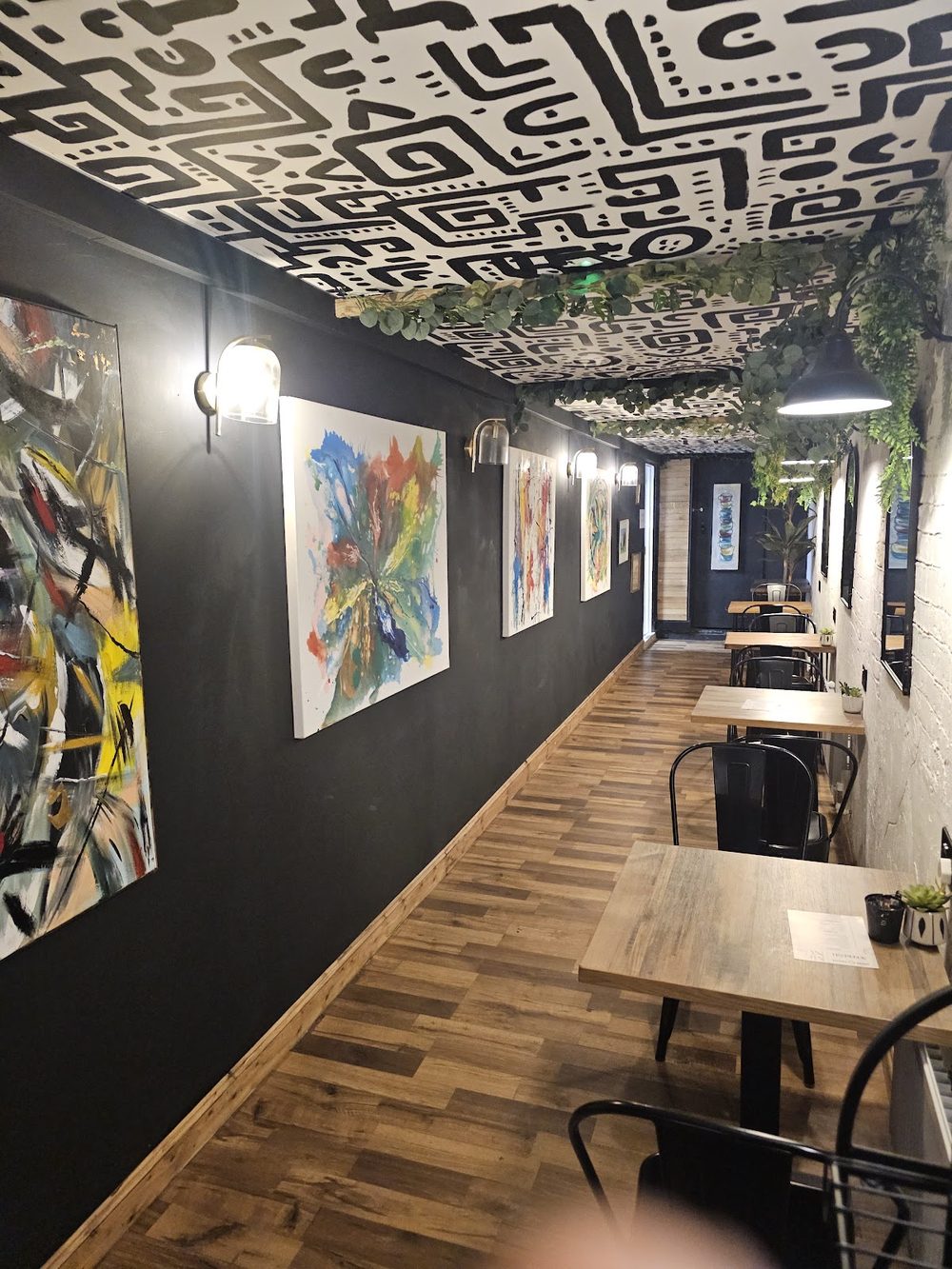 A narrow room lined with colourful framed artwork on a black wall, a patterned ceiling above a row of tables